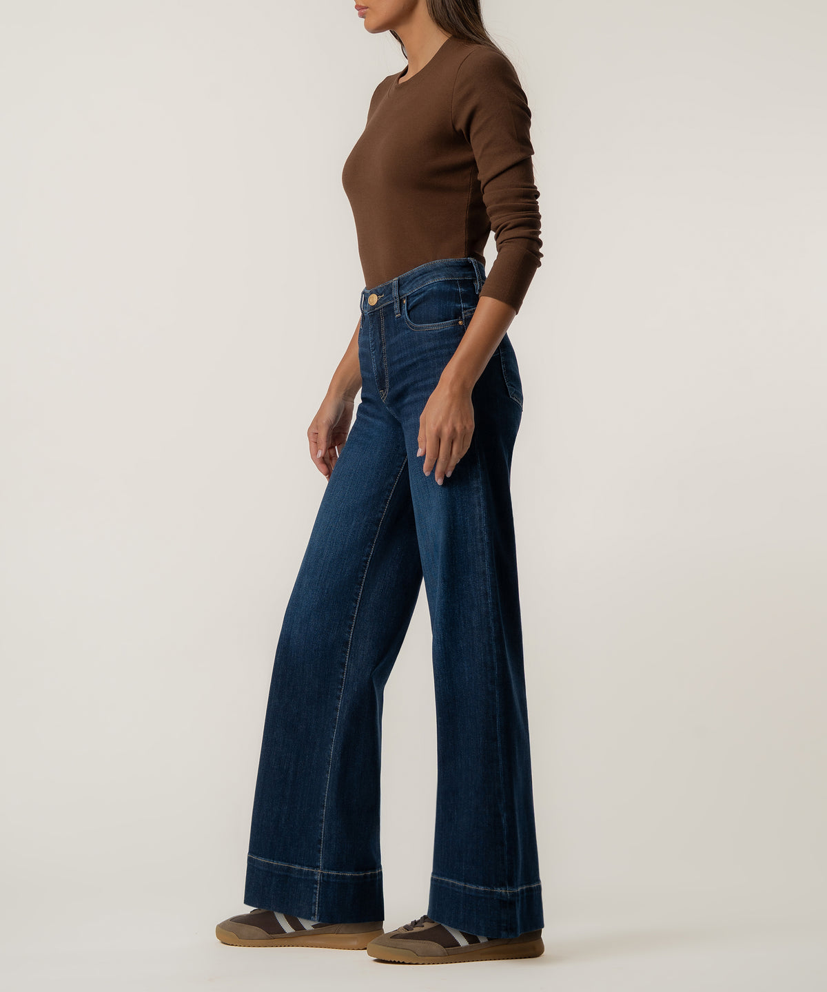 A woman in a brown long-sleeve top wears high-rise dark blue wide-leg jeans with wide cuffed hems and brown sneakers.