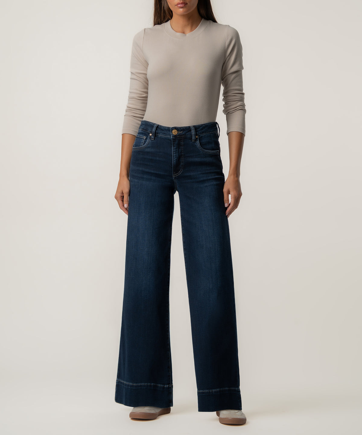 A woman stands wearing dark blue high-rise wide-leg jeans with wide hems, a fitted beige long-sleeve top, and light sneakers.