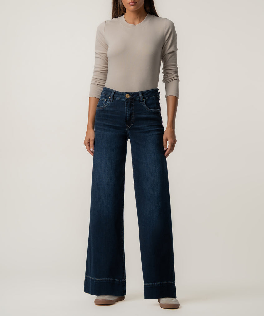 A woman stands wearing dark blue high-rise wide-leg jeans with wide hems, a fitted beige long-sleeve top, and light sneakers.