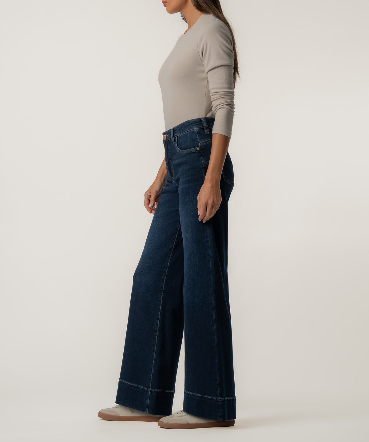 A woman stands in profile wearing high-rise dark blue wide-leg jeans, a fitted gray long-sleeve top, and cream sneakers.