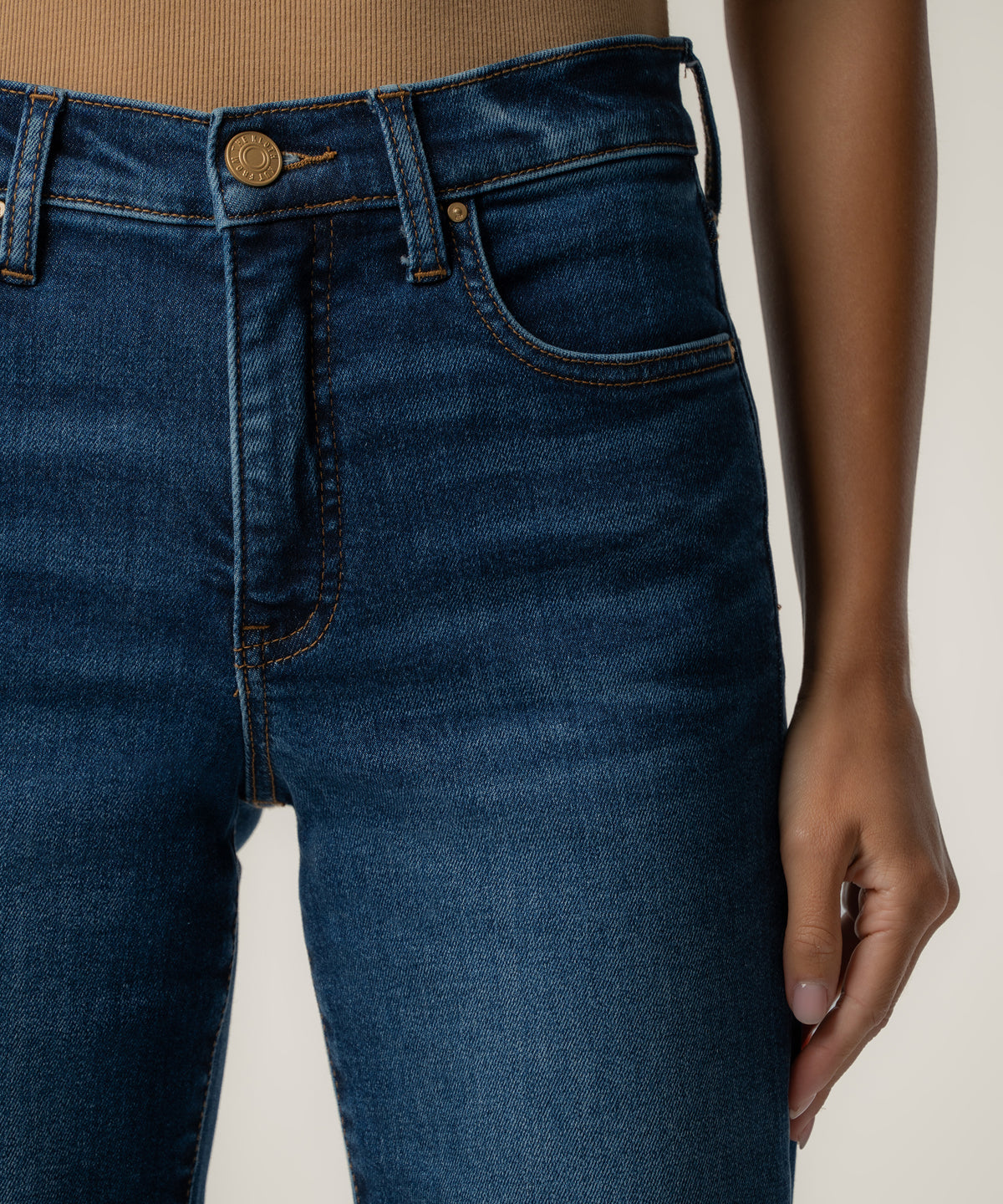Close-up of a person wearing high-rise dark blue jeans with a brass button, orange topstitching, and a tan ribbed top.