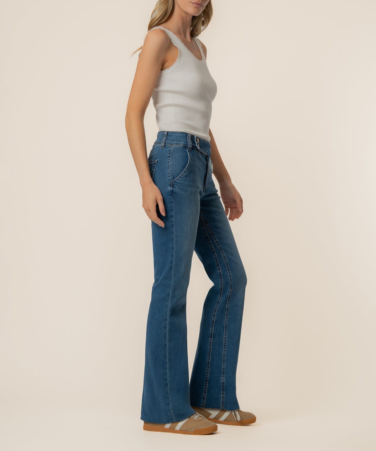 Woman wearing high-rise medium-wash flare jeans with a white ribbed tank top and tan striped sneakers.