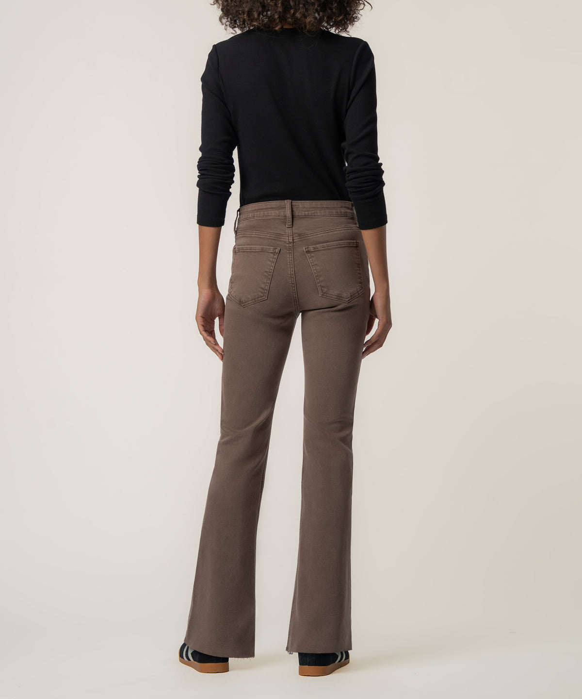 Back view of a woman wearing mocha brown high rise flare pants with raw hems, a black long-sleeve top, and sneakers.