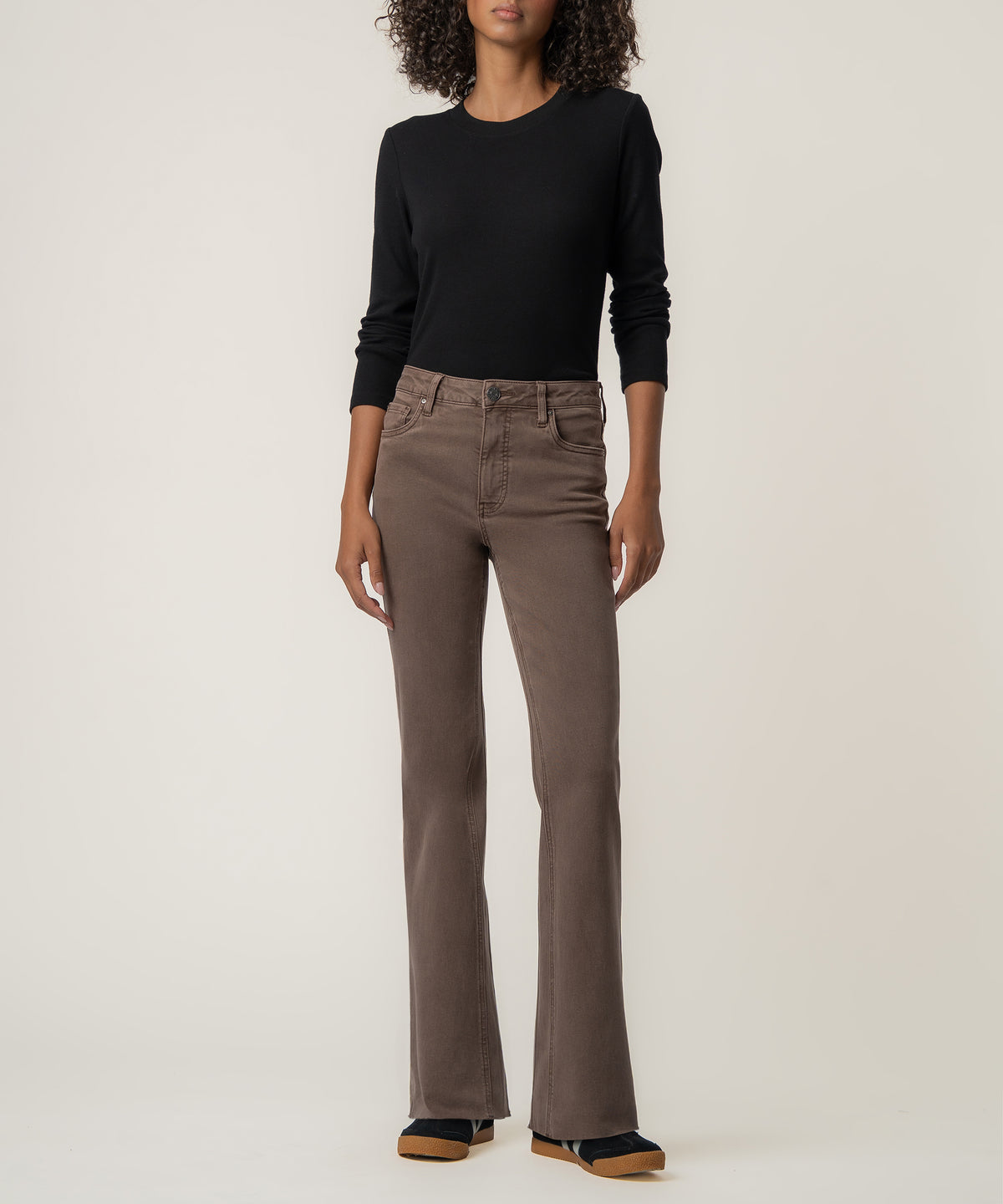 A woman wears high-rise brown flared pants with a raw hem, a black long-sleeve top, and dark sneakers.