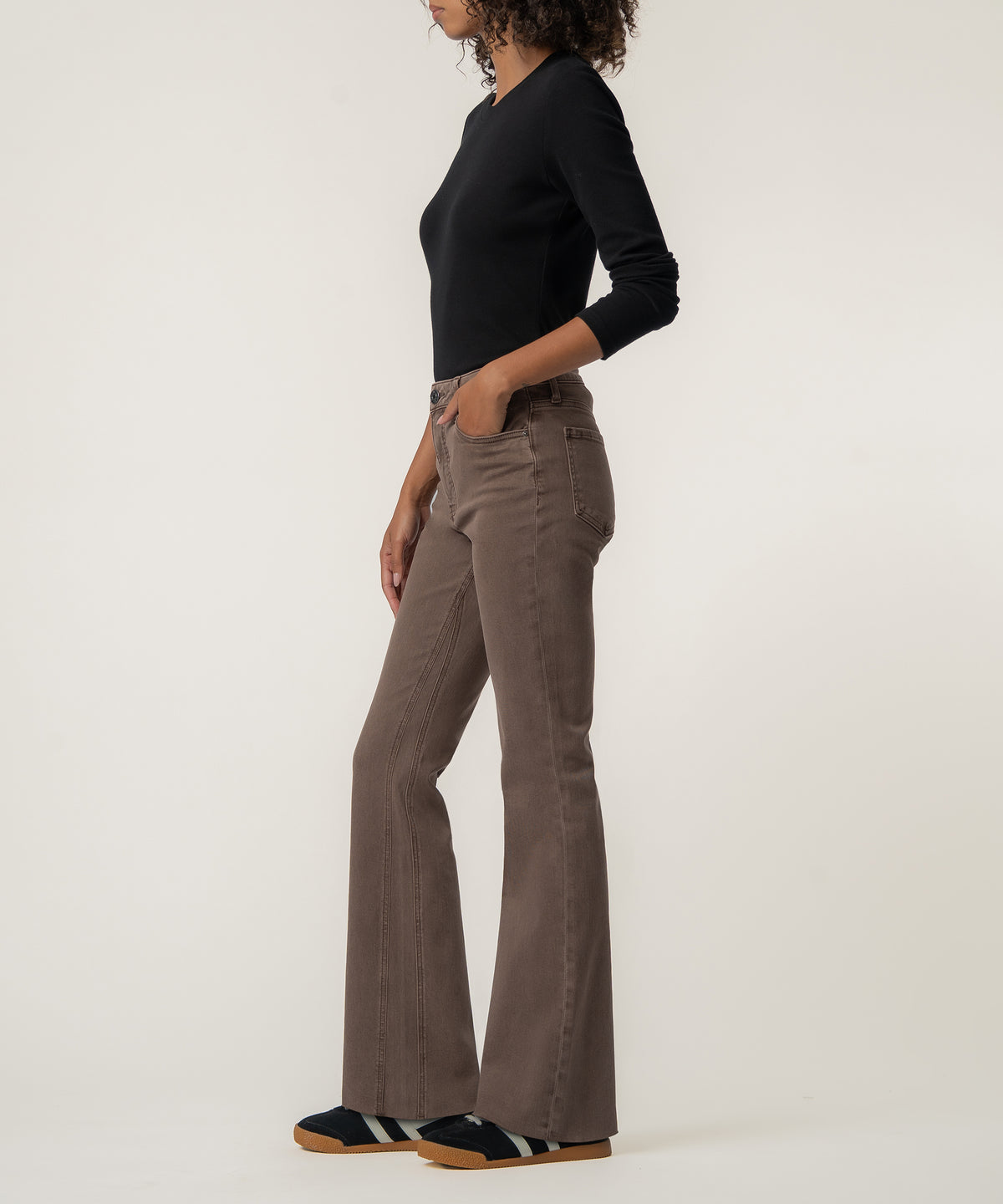 A woman stands in profile wearing high-rise brown flare pants, a fitted black long-sleeve top, and dark sneakers with gum soles.
