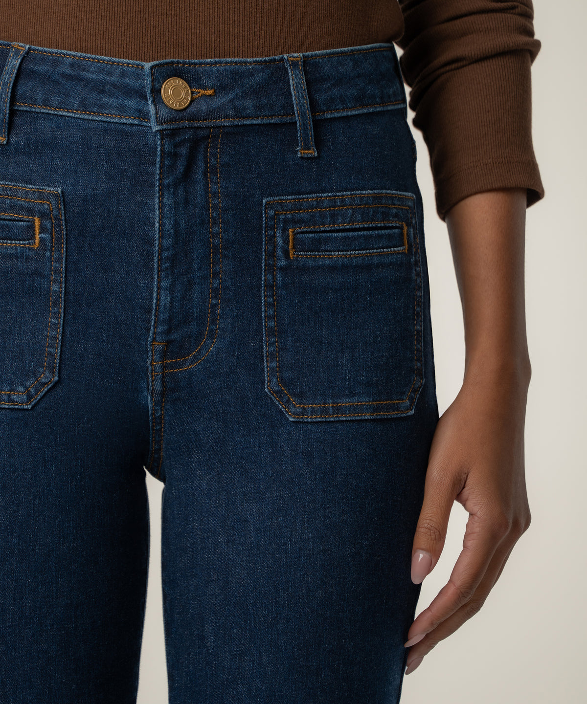 Close-up of a person wearing dark blue high-rise flare jeans with square front patch pockets, orange topstitching, and a brass button.
