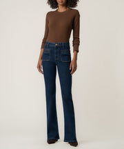 A model stands wearing high-rise dark blue flare jeans with front patch pockets, a brown long-sleeve top, and dark pointed shoes.