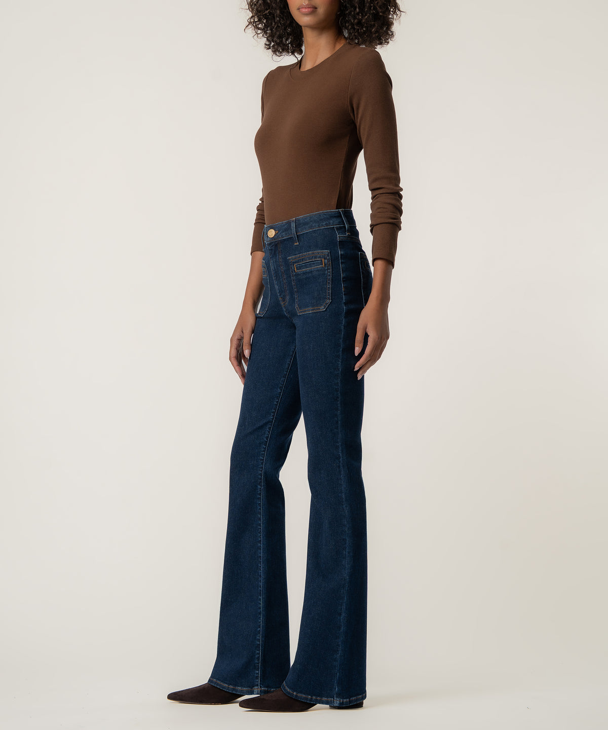 A woman models dark blue high-rise flare jeans with front patch pockets, a brown long-sleeve top, and dark pointed shoes.