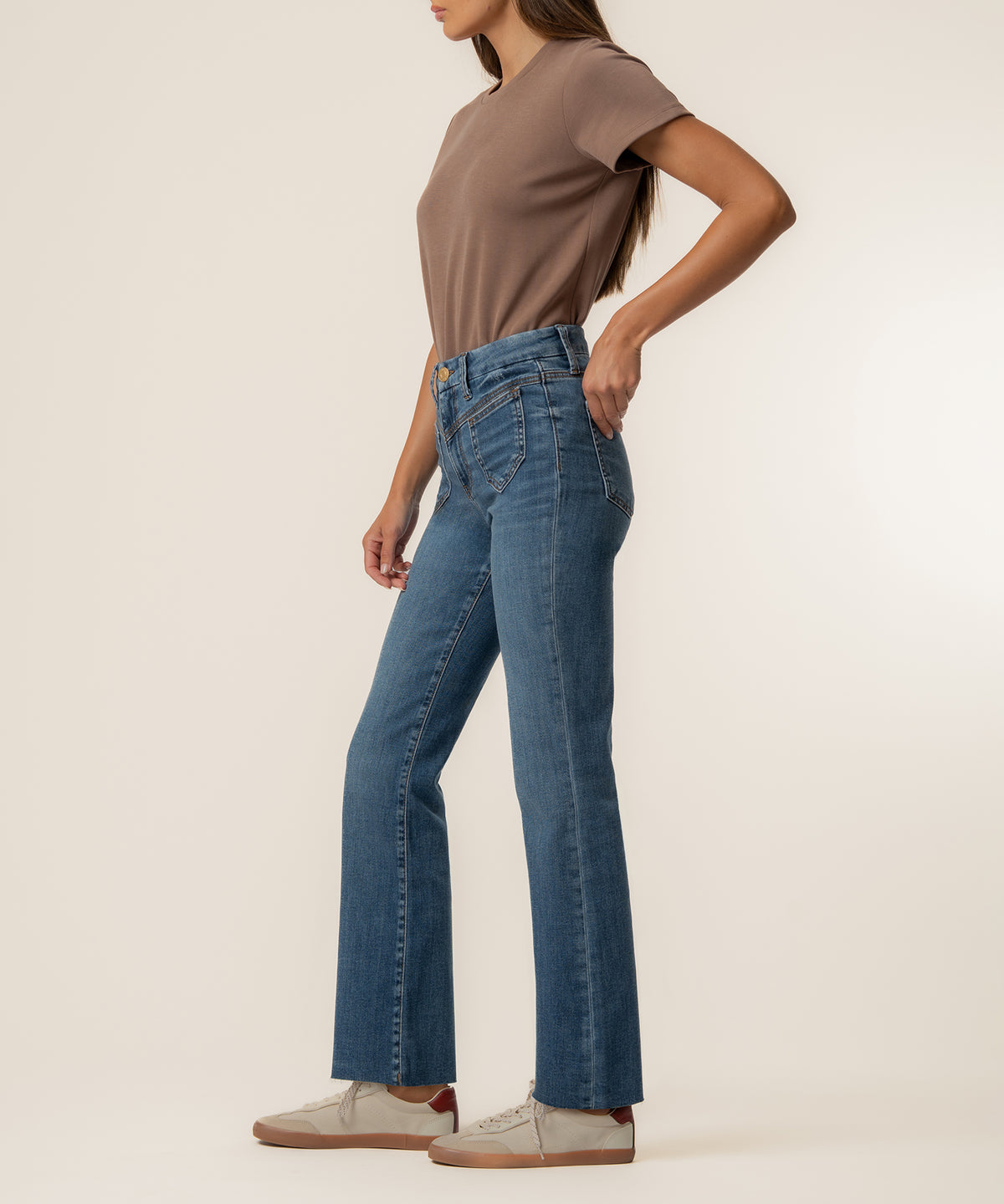 Woman wearing high-rise ankle flare jeans in medium blue denim with a taupe t-shirt and cream sneakers.