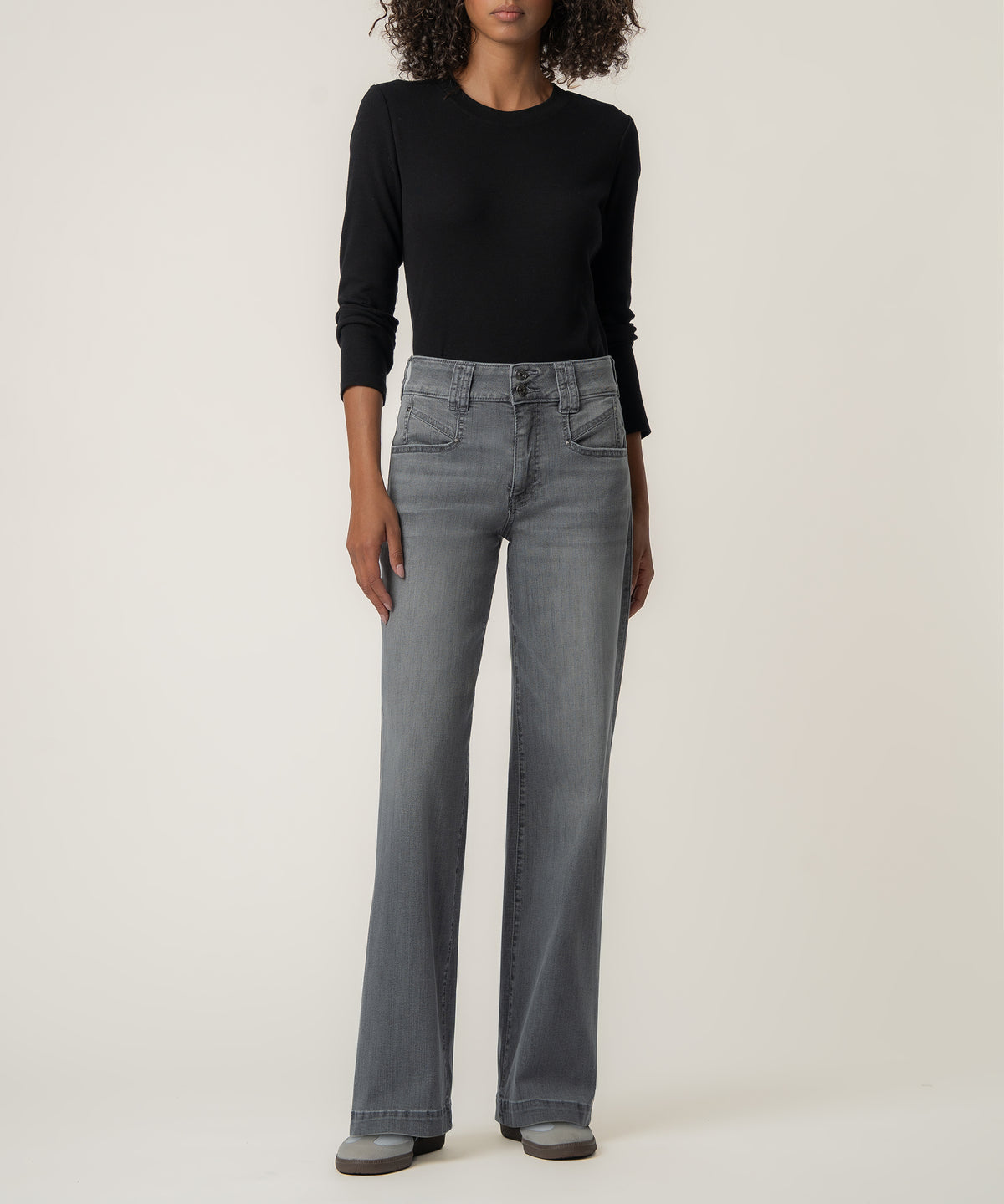 A woman with curly hair wears high-rise gray wide-leg jeans with a black long-sleeve top and gray sneakers.
