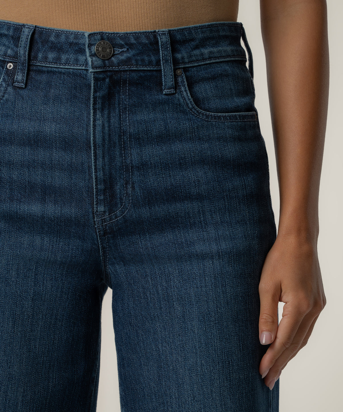 Close-up of a person wearing dark blue high-rise wide-leg jeans with a button closure, belt loops, and a tan top.