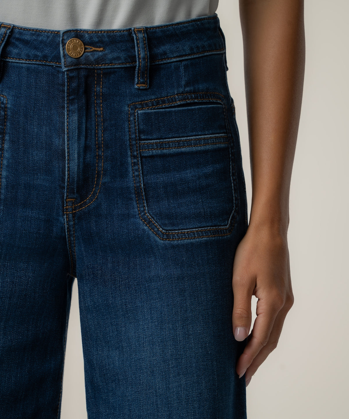 Close-up of a person wearing high-rise dark blue wide-leg jeans with a gold button, orange topstitching, and a large patch pocket.