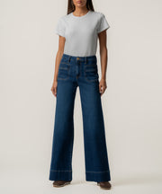 A woman wears high-rise wide-leg dark blue jeans with front patch pockets, a light gray t-shirt, and brown shoes.