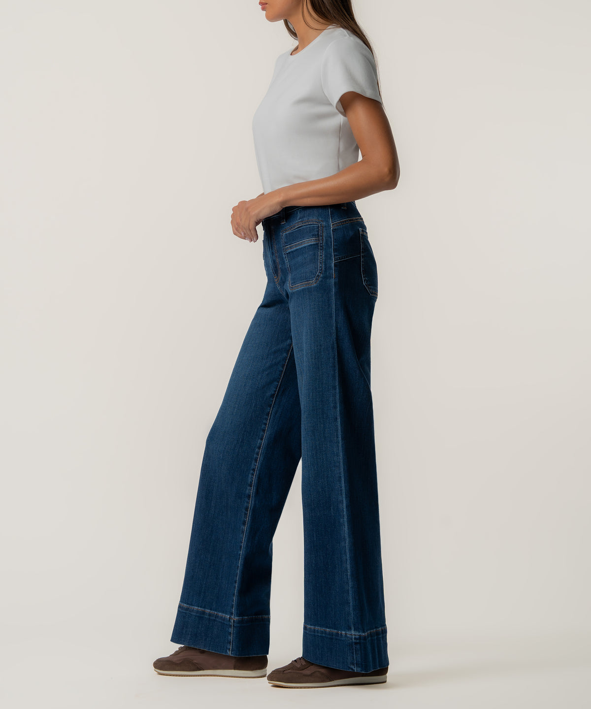 A woman stands in profile wearing high-rise wide-leg dark blue jeans with front patch pockets, a white t-shirt, and brown sneakers.