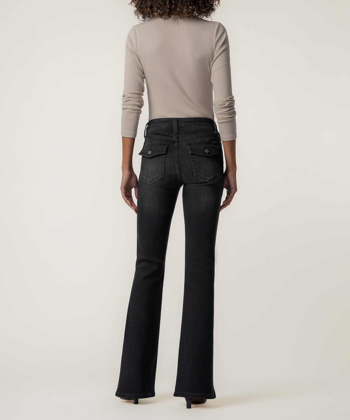 Back view of a woman wearing faded black high rise flare jeans with button flap back pockets, a beige long sleeve top, and heels.