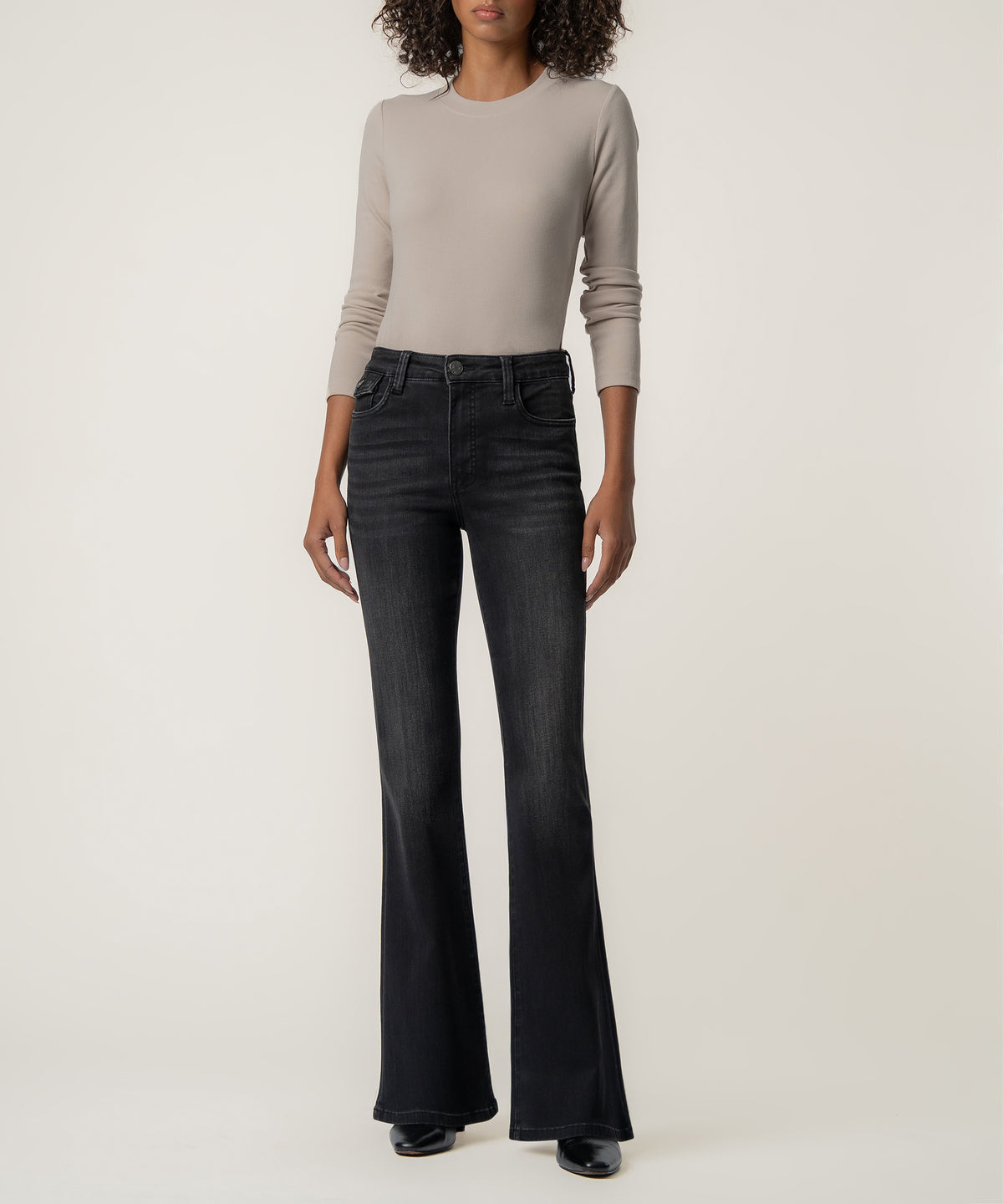 A woman stands facing forward in faded black high-rise flare jeans, a taupe long-sleeve top, and black pointed boots.