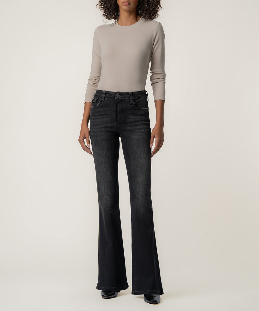 A woman stands facing forward in faded black high-rise flare jeans, a taupe long-sleeve top, and black pointed boots.