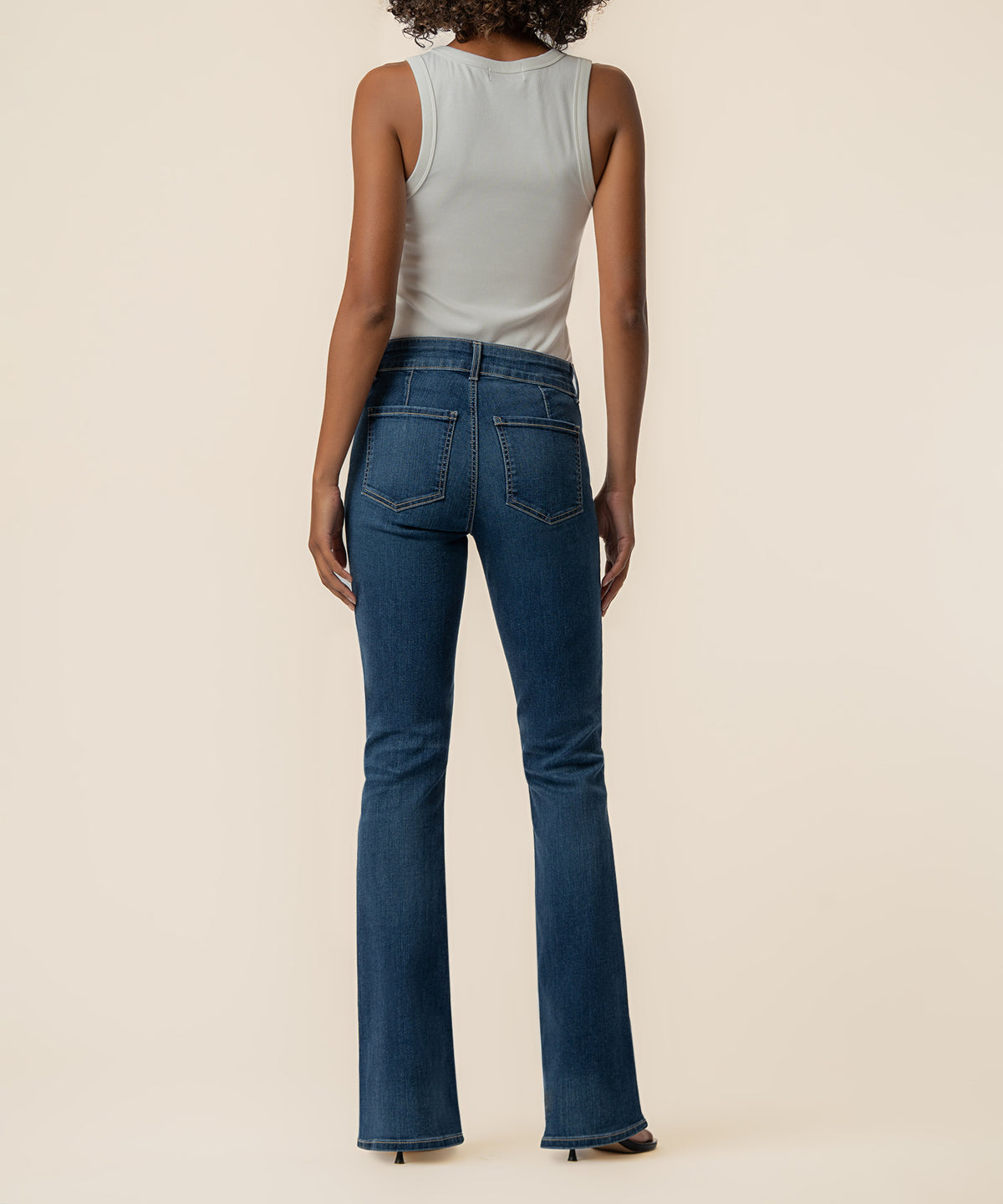Woman wearing high-rise dark blue bootcut jeans with flared hem and white sleeveless tank top, rear view.