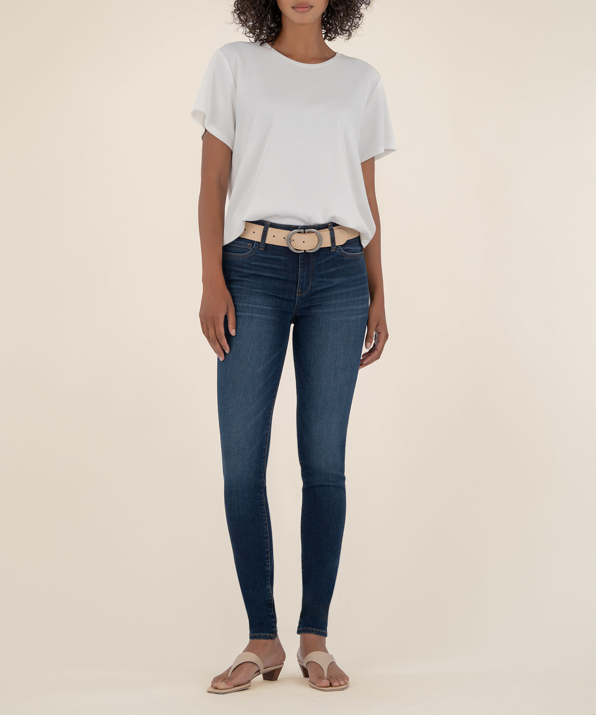 Woman wearing white t-shirt, dark blue skinny jeans with tan belt, and tan slide sandals.