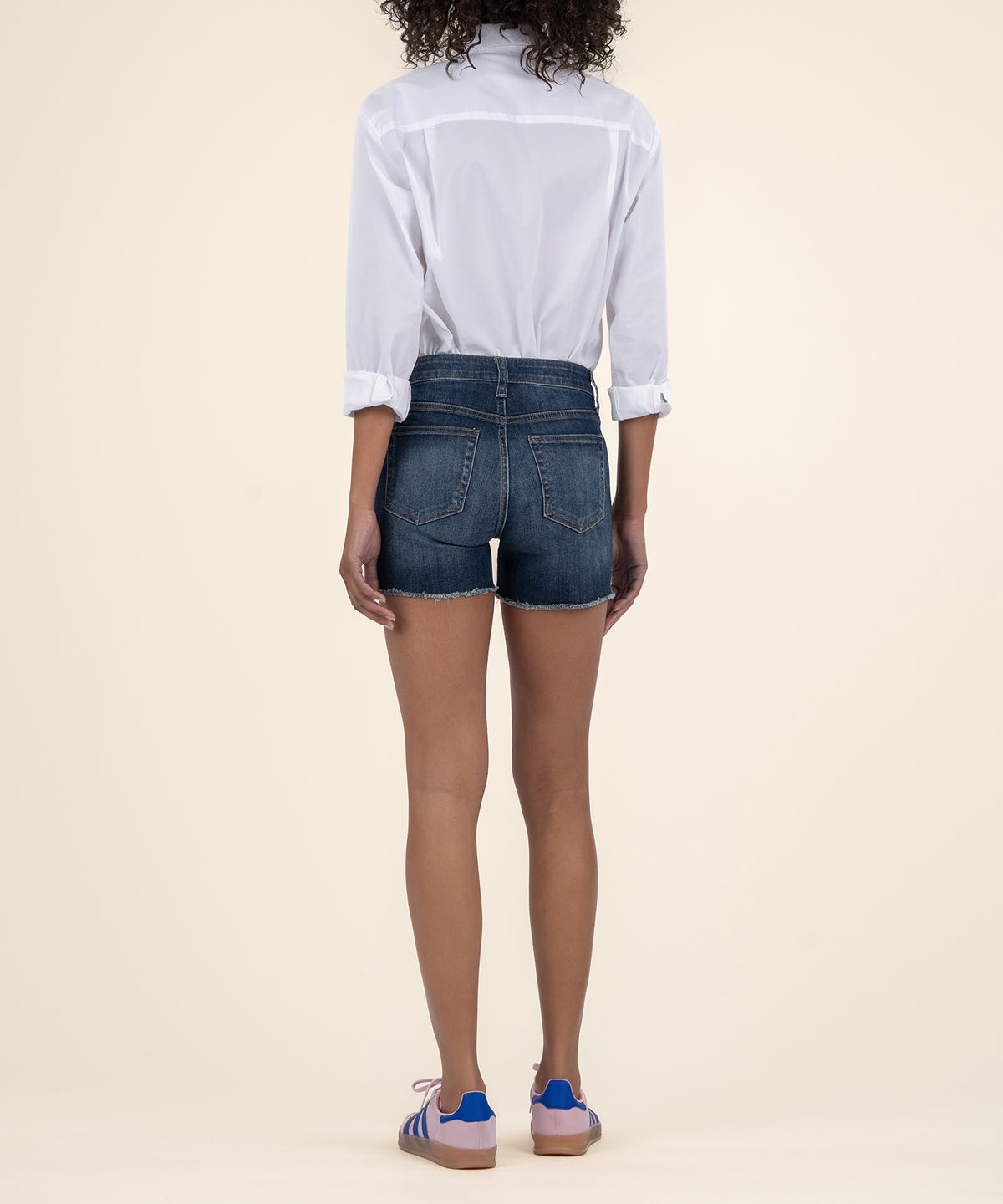 Woman wearing white button-up shirt and dark wash denim shorts with frayed hem, back view.