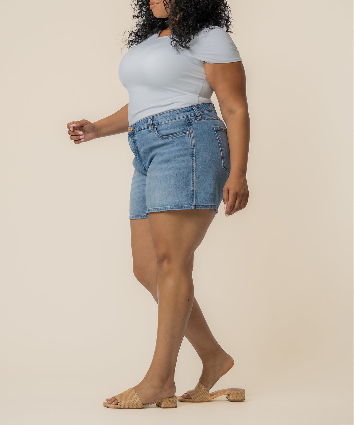 Light-wash denim high-rise shorts on model paired with white off-shoulder t-shirt and tan slides.