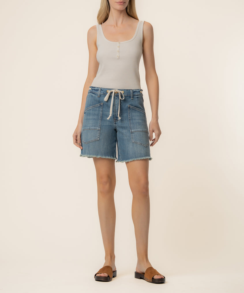 Woman wearing a light blue denim high-rise short with cargo pockets and frayed hem, paired with a white tank top.
