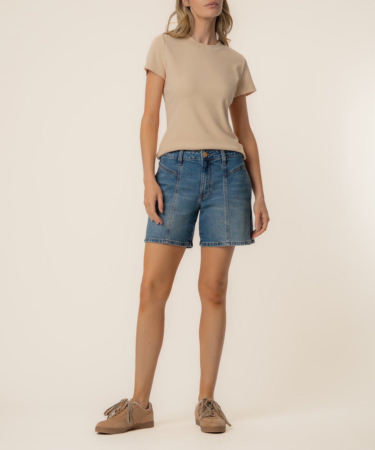 Woman wearing a beige t-shirt and blue denim high-rise shorts with a glossy finish and brown lace-up shoes.