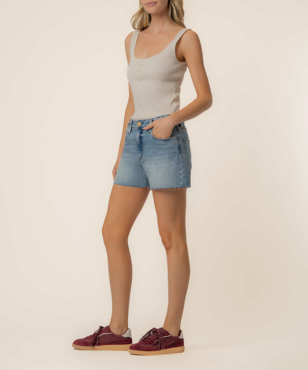 Woman wearing high-rise denim shorts in light blue wash with frayed hem and cream tank top.