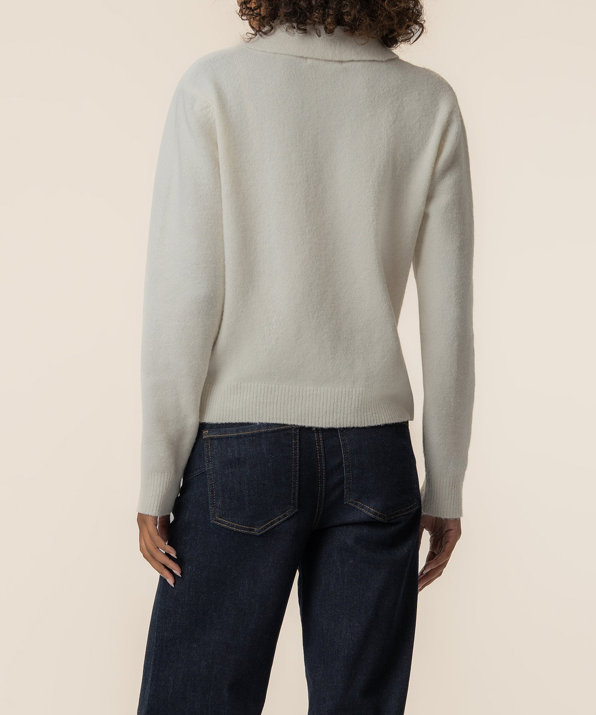 Cream knit sweater with long sleeves and ribbed trim worn with dark denim jeans, shown from back view.