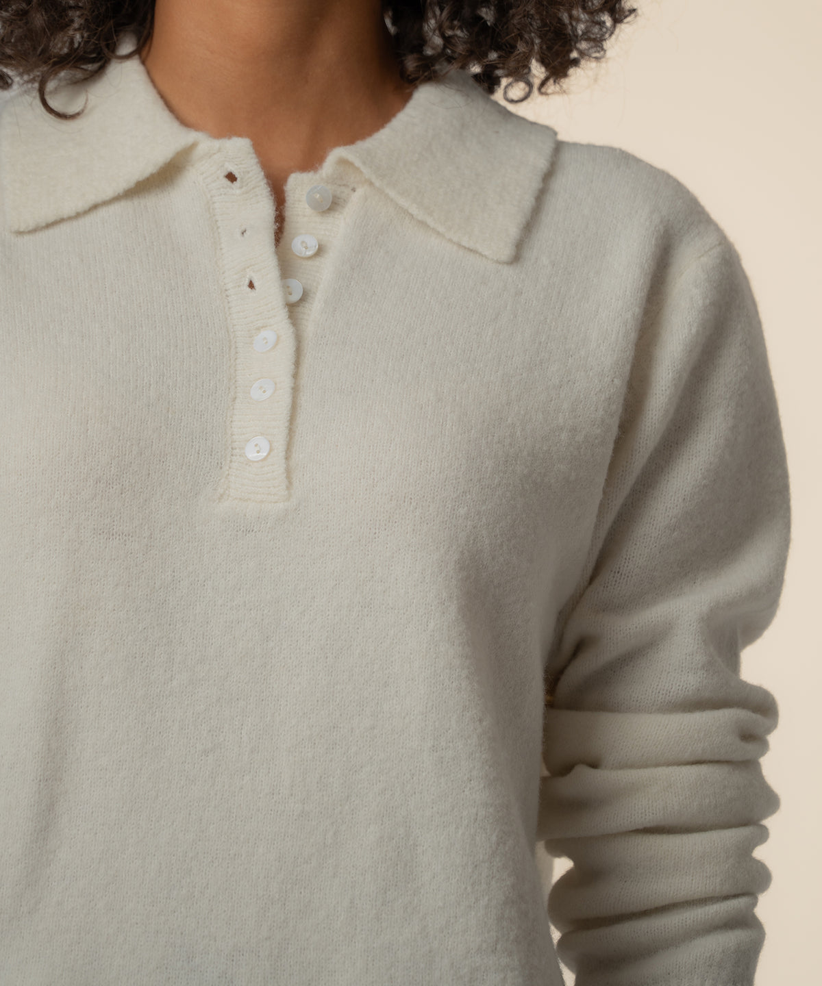 Cream-colored sweater with collared neckline and white button placket front, shown on model with dark curly hair.