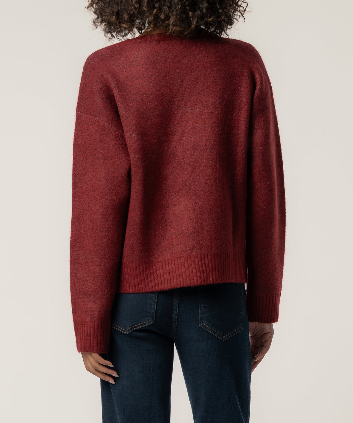 Back view of a person with curly hair wearing a dark red knit cardigan with ribbed hem and cuffs, paired with dark jeans.