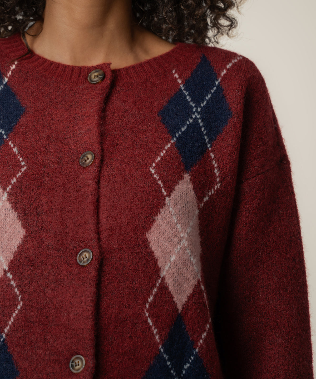 Close-up of a person wearing a dark red knit cardigan with navy and pink argyle diamonds and brown buttons.