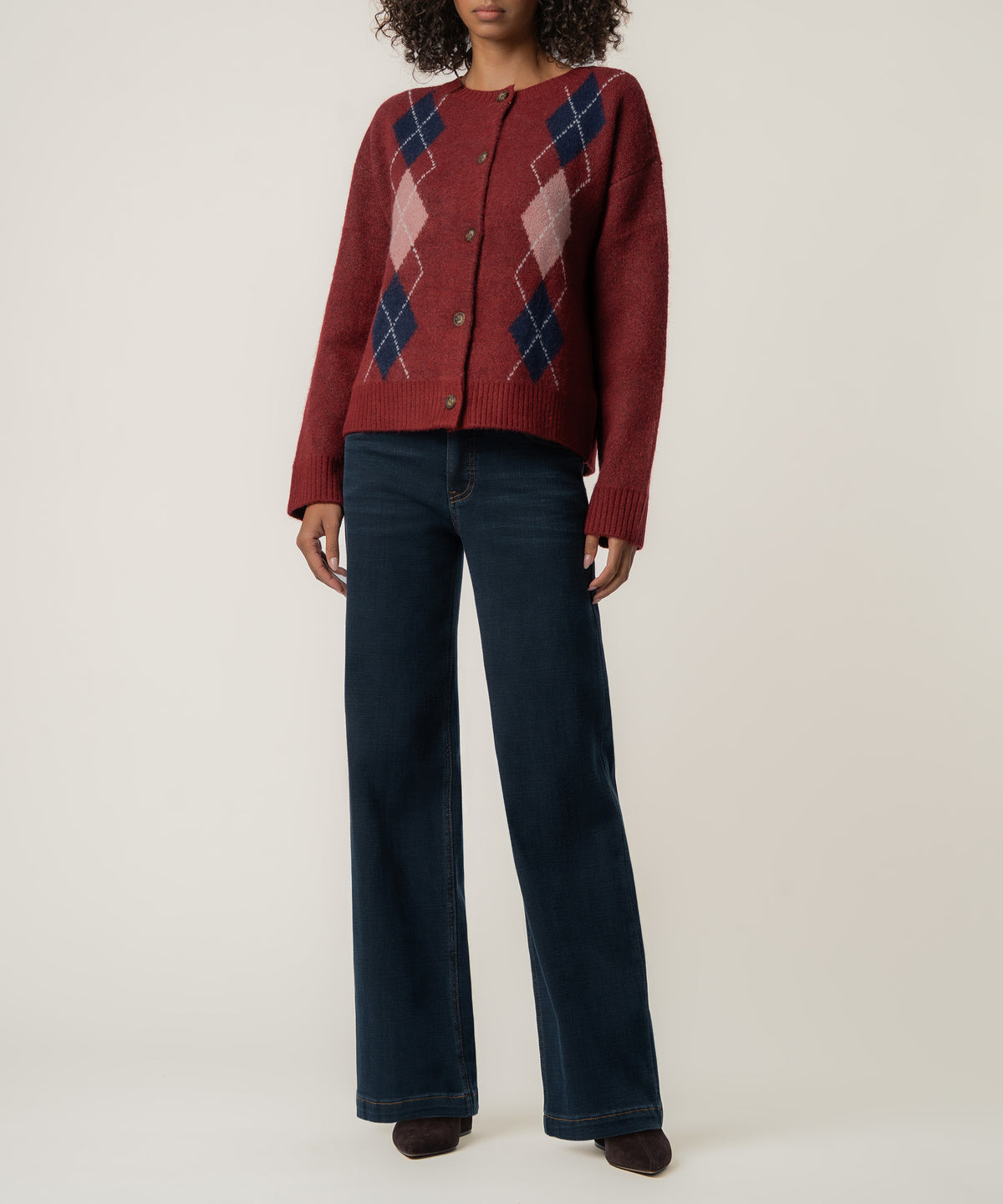 A woman wears a dark red buttoned cardigan with navy and pink argyle diamonds, paired with wide-leg dark jeans.