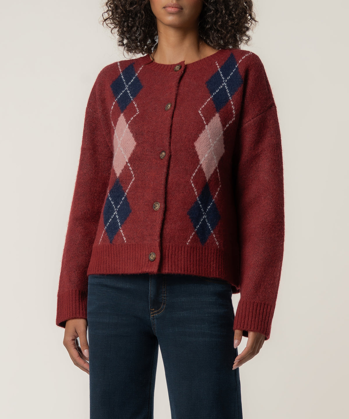 A woman wears a dark red button-front cardigan with navy and pink argyle diamonds, paired with dark blue jeans.