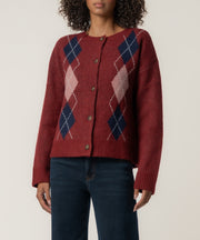A woman wears a dark red button-front cardigan with navy and pink argyle diamonds, paired with dark blue jeans.