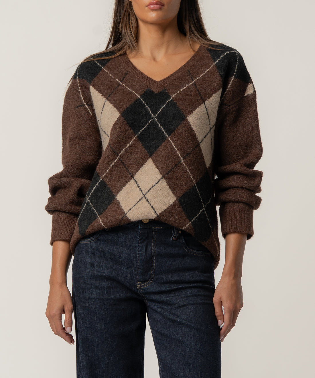 Woman wearing a brown argyle V-neck sweater with black and cream diamond pattern and ribbed cuffs, paired with dark jeans.