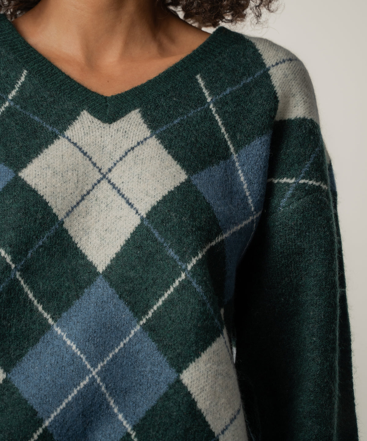 Close view of a person wearing a dark green V-neck sweater with a gray and blue argyle diamond pattern and thin white lines.