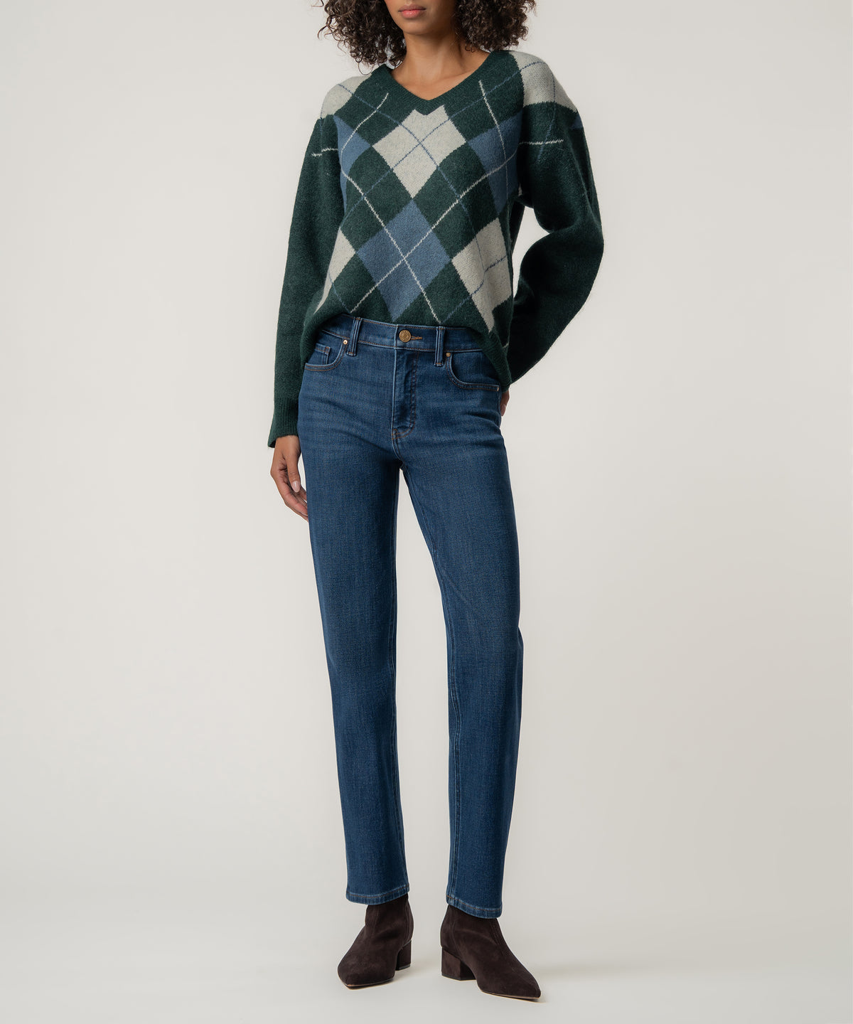 A woman wears a dark green V-neck sweater with a blue and gray argyle pattern, blue jeans, and brown suede ankle boots.
