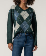 A woman wears a dark green V-neck sweater with a blue and cream argyle pattern, paired with high-waisted blue jeans.