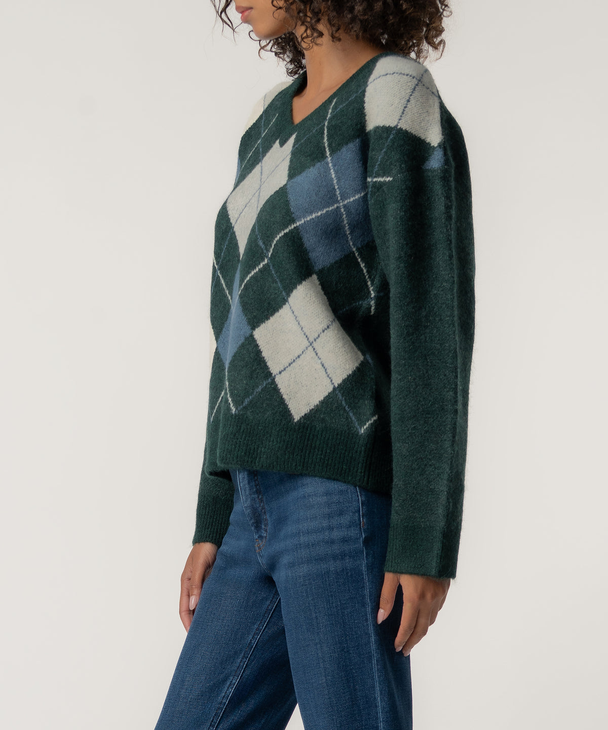 A woman in profile wears a dark green V-neck sweater with cream and blue argyle diamonds, paired with blue jeans.