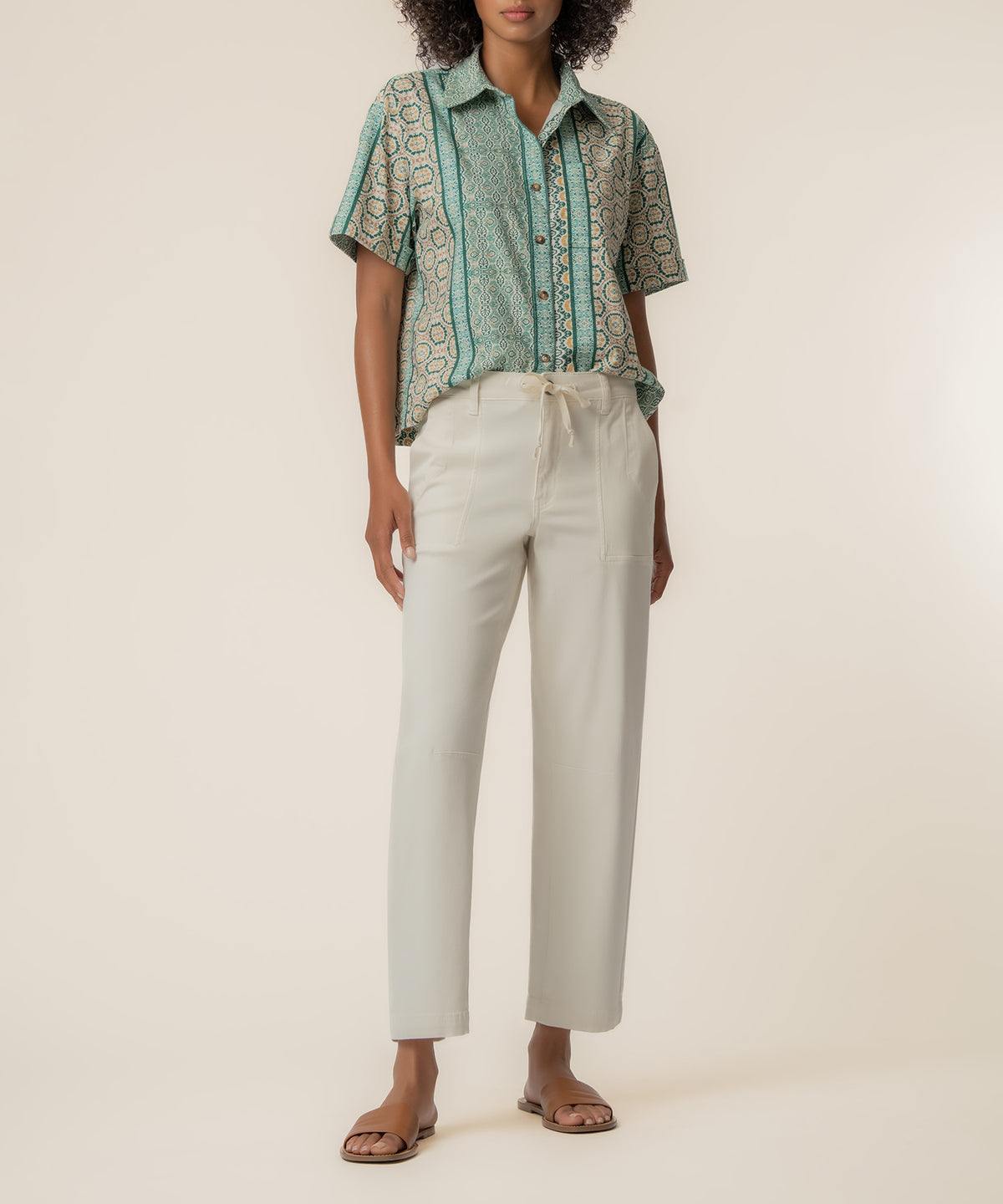 Woman wearing short-sleeved patterned linen shirt in sage green with cream trousers and brown sandals.
