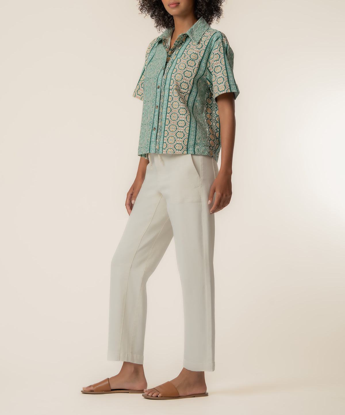 Woman wearing a short-sleeved button-up linen top with geometric patterning in teal and cream tones, paired with cream-colore