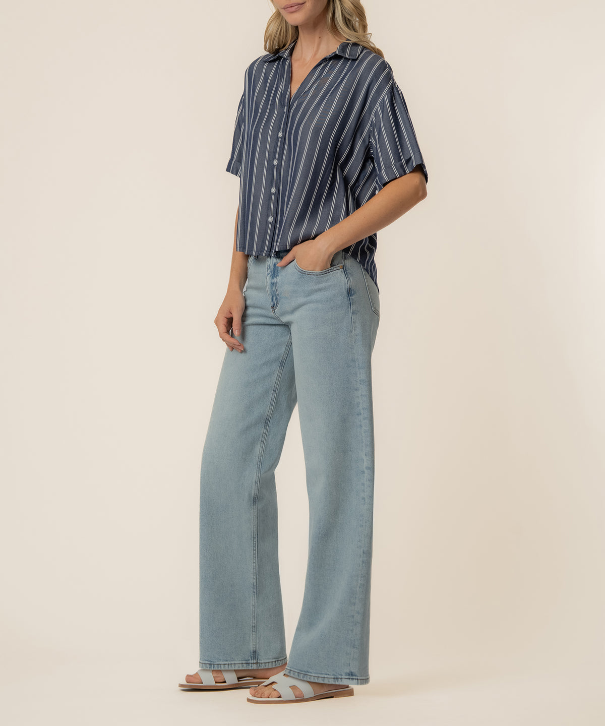 Woman wearing indigo and white striped boxy button-up shirt with light blue jeans and tan sandals.