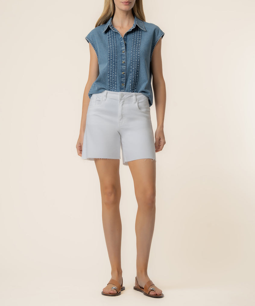 Woman wearing light blue denim sleeveless shirt with vertical pintucks and white denim shorts with raw hem.